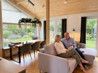Evergreen Wohnbereich mit einem Paar auf dem grauen Sofa, Holztisch und Blick in den Garten